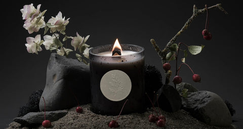 a fischersund scented candle in its blackened galss jar and burning within a crafted landscape that is both clearly of iceland with its dark black sand and rocks, twigs, and white foliage but also otherworldly and spiritual