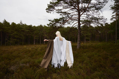 lady in a field on a forest edge with various throws and blankets over her arms