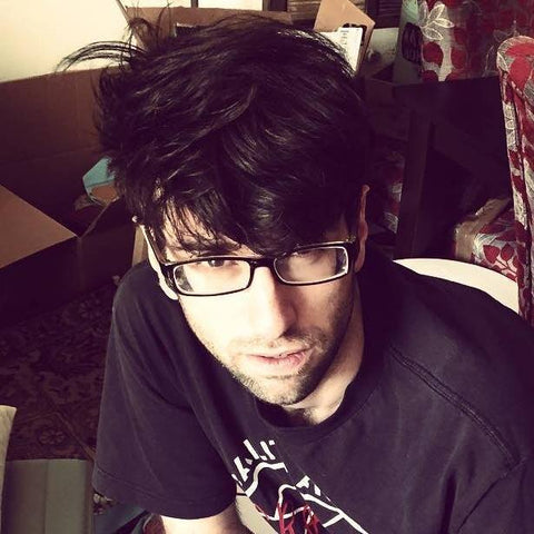 A photograph of co-author of The Book of the Magical Mythical Unicorn, Alfonso Colasuonno. Alfonso is wearing eyeglasses and a t-shirt of the anarcho-punk band Crass amidst a messy living room.