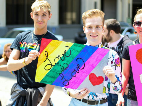 Two gay blond men at a Pride Parade holding a rainbow-colored sign that says "Love is Love"