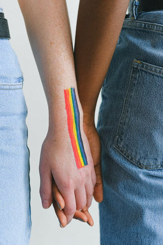 Two women holding hands, one of whom has a rainbow pride flag painted on her hand to symbolize the LGBTQI+ community