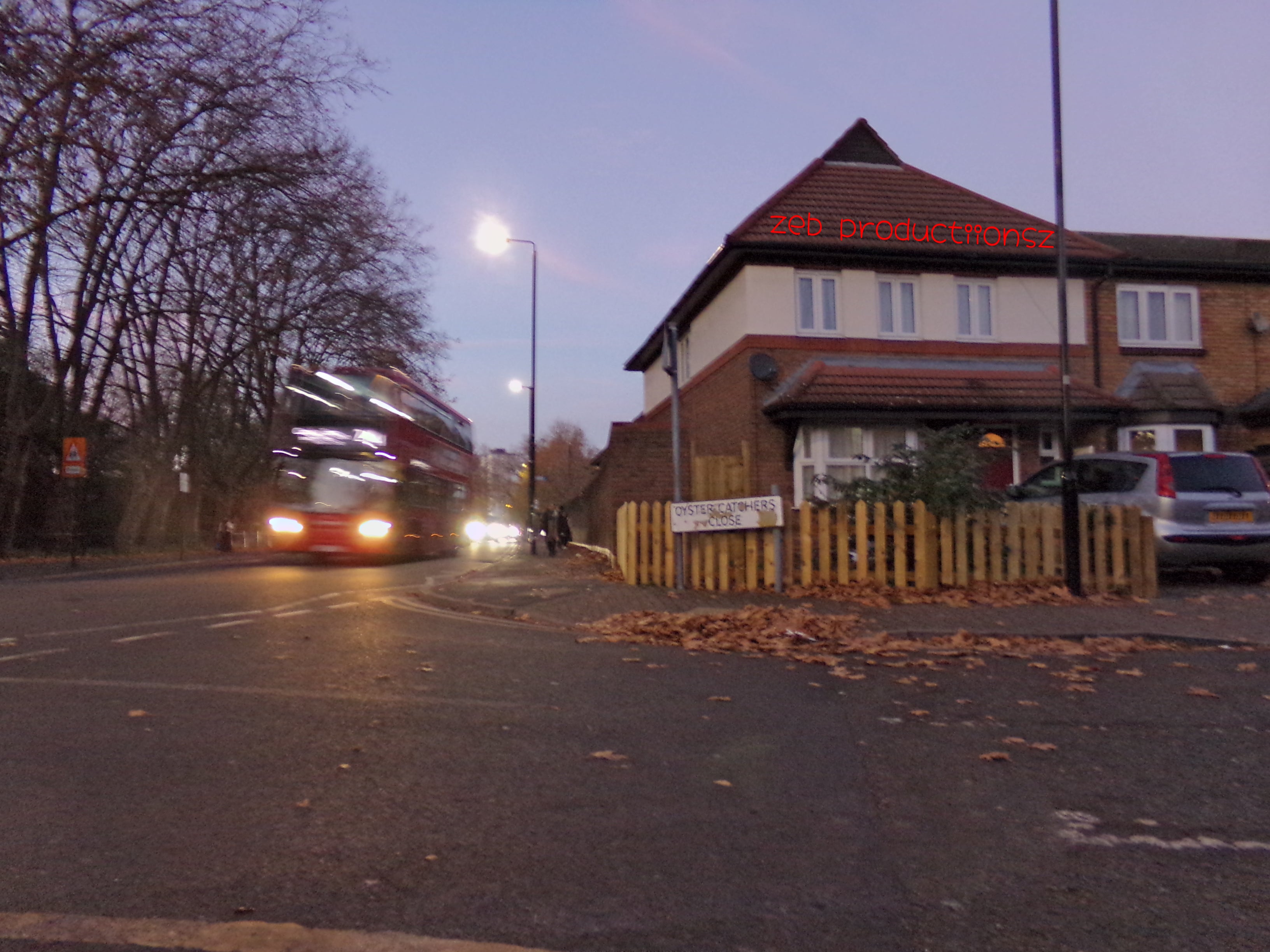 A wide frame picture of the end of a close, there are bare trees on the left of the picture, a red bus driving pass. the pavement is empty. the house on the right is brown and white, there is a silver jeep style car in the drive way. on the roof it says "zeb productiionzs" in a red graffiti style