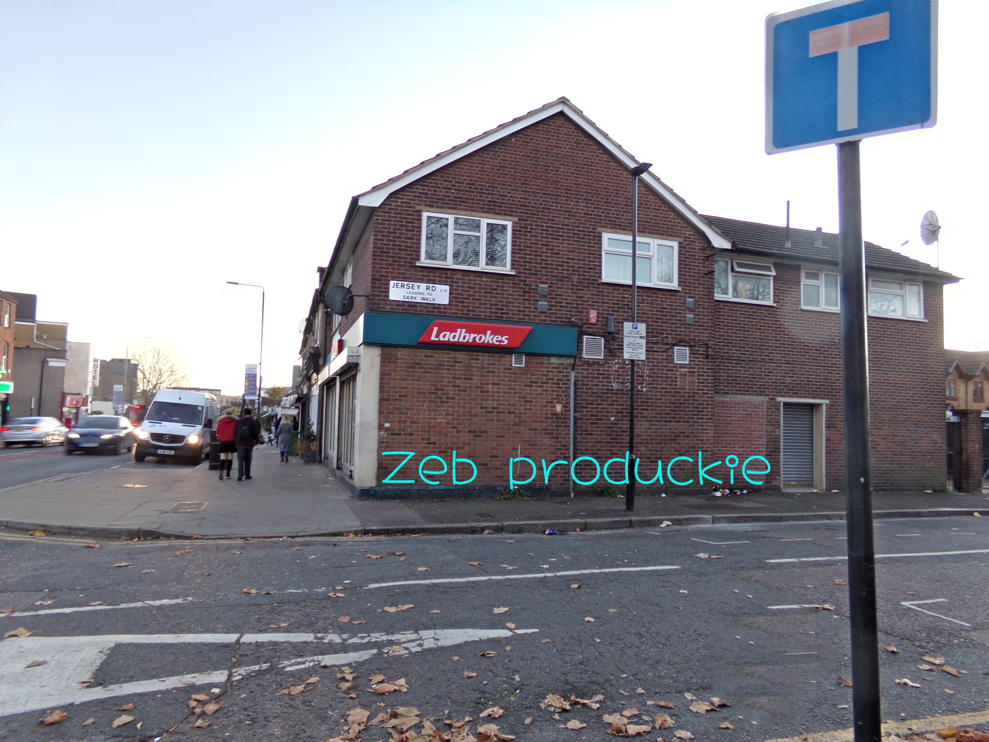 A picture of a side road in East London. There is a sign on the left showing a dead end. There is a brown brick building with a rectangular teal and red sign, white text reads "Ladbrokes". Theres a busy street in the background. There are brown leaves on the ground.