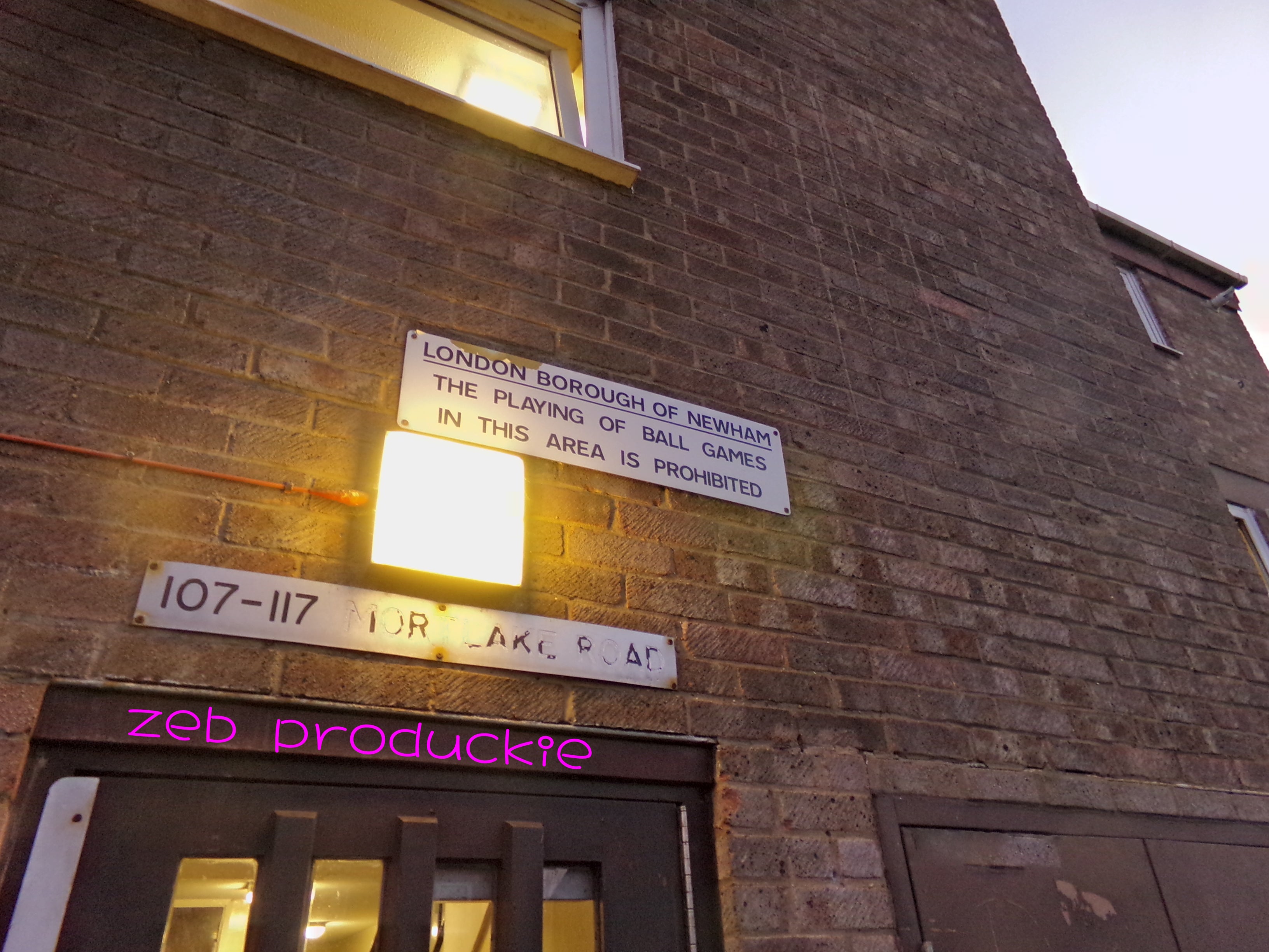 It is a close up of a brown building. On the corner left, a maroon door is shown, above is silver metal sign with the numbers and words faded. There is a square fluorescent light directly above. To the right of the light there is a silver metal sign that reads "The London Borough of Newham - The playing of ball games in this area is prohibited." A corner of a window with yellow light is shown an the top left corner of the photo