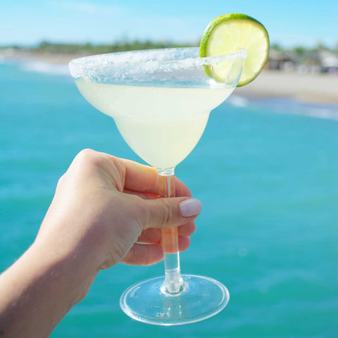 Feminine hands holding a Margarita cocktail with lime wheel garnish against an ocean background 