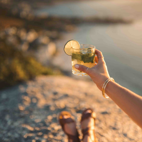 Woman holding up a Mojito cocktail on a rocks glass against a beach background