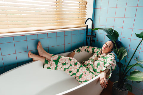 A model is laying down in an empty white bathtub wearing plastic-free loungewear in a whimsical green clouds print