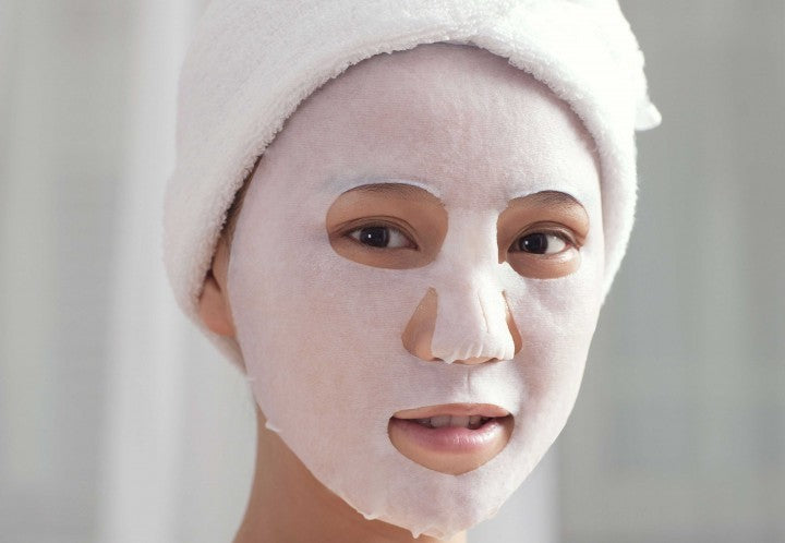 A young woman of Asian descent with a well applied sheet mask on her face. Her hair is wrapped up in a towel.