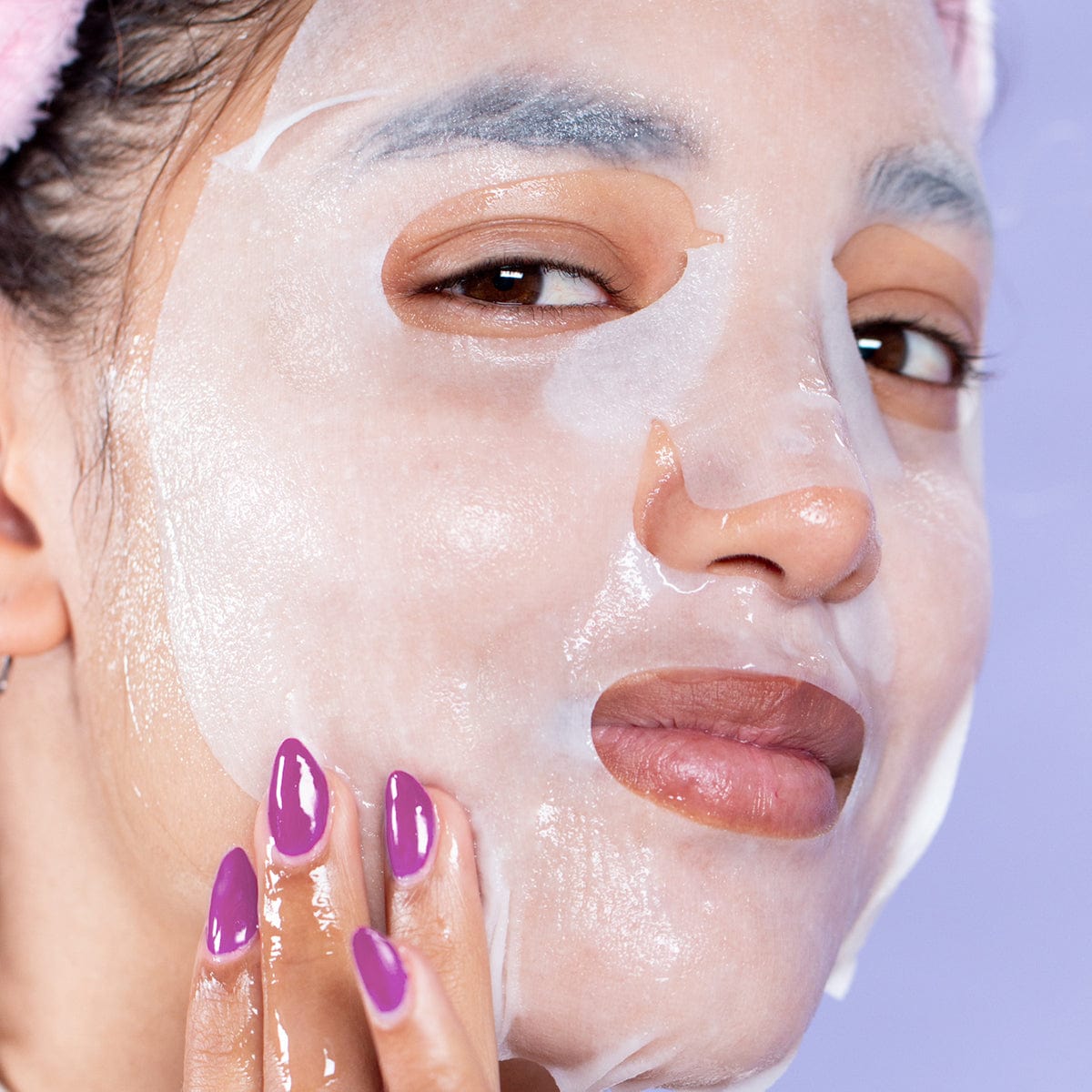 Woman of Asian heritage with a sheet mask on her face. She has light purple nails, and her hair is pulled back