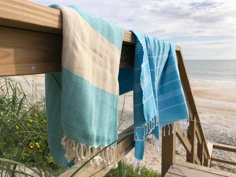Two Turkish towels with different shades of blue on a railing with a beach and the ocean behind them