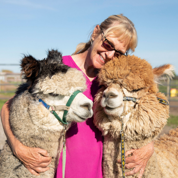 woman smiling with her arms wrapped around two alpacas