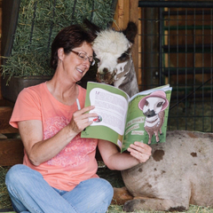 a woman sitting in a barn reading to an alpaca