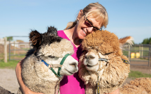 woman smiling standing outside with her arms wrapped around two alpacas