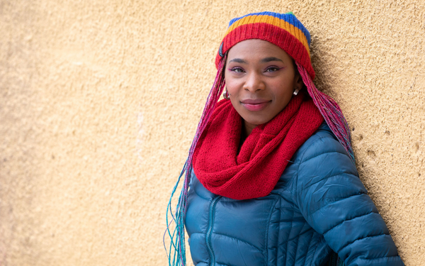 black woman leaning against a tan wall wearing rainbow beartooth beanie and red handmade alpaca wool infinity scarf