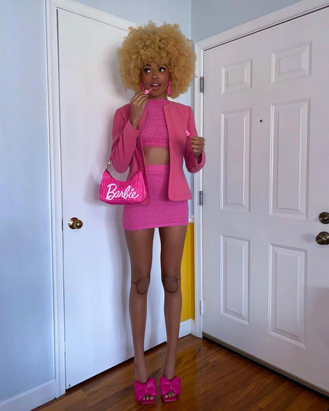 A woman wears a DIY Barbie Halloween costume, featuring a matching pink jacket, crop top, and mini skirt, with hot pink high heels and purse.