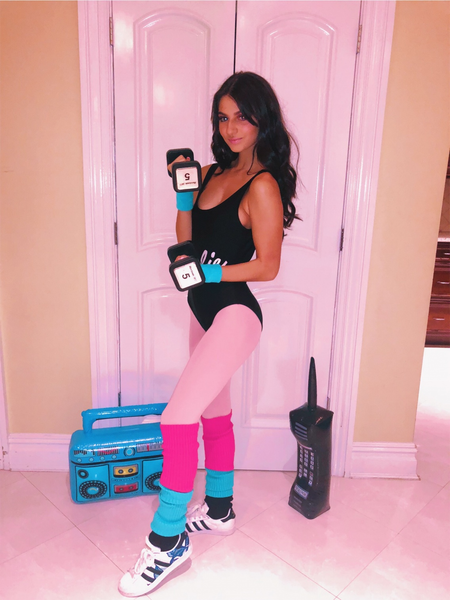 A woman wears a DIY Barbie Halloween costume, featuring a black bodysuit, hot pink leggings, and pink and blue legwarmers, while she lifts weights.