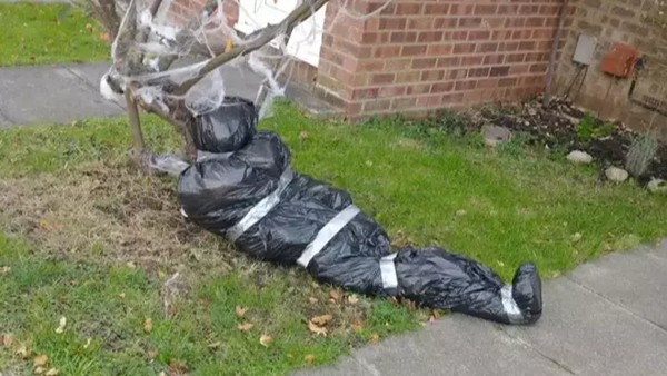 A body bag Halloween decoration sits propped against a tree.