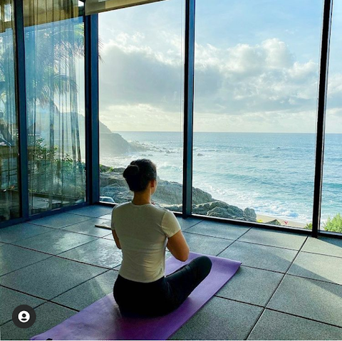 Dr. Mamina sits on a yoga mat, crosses her legs, and put her hands together in a praying, meditating position. Behind a window, she is onlooking the beach shore.