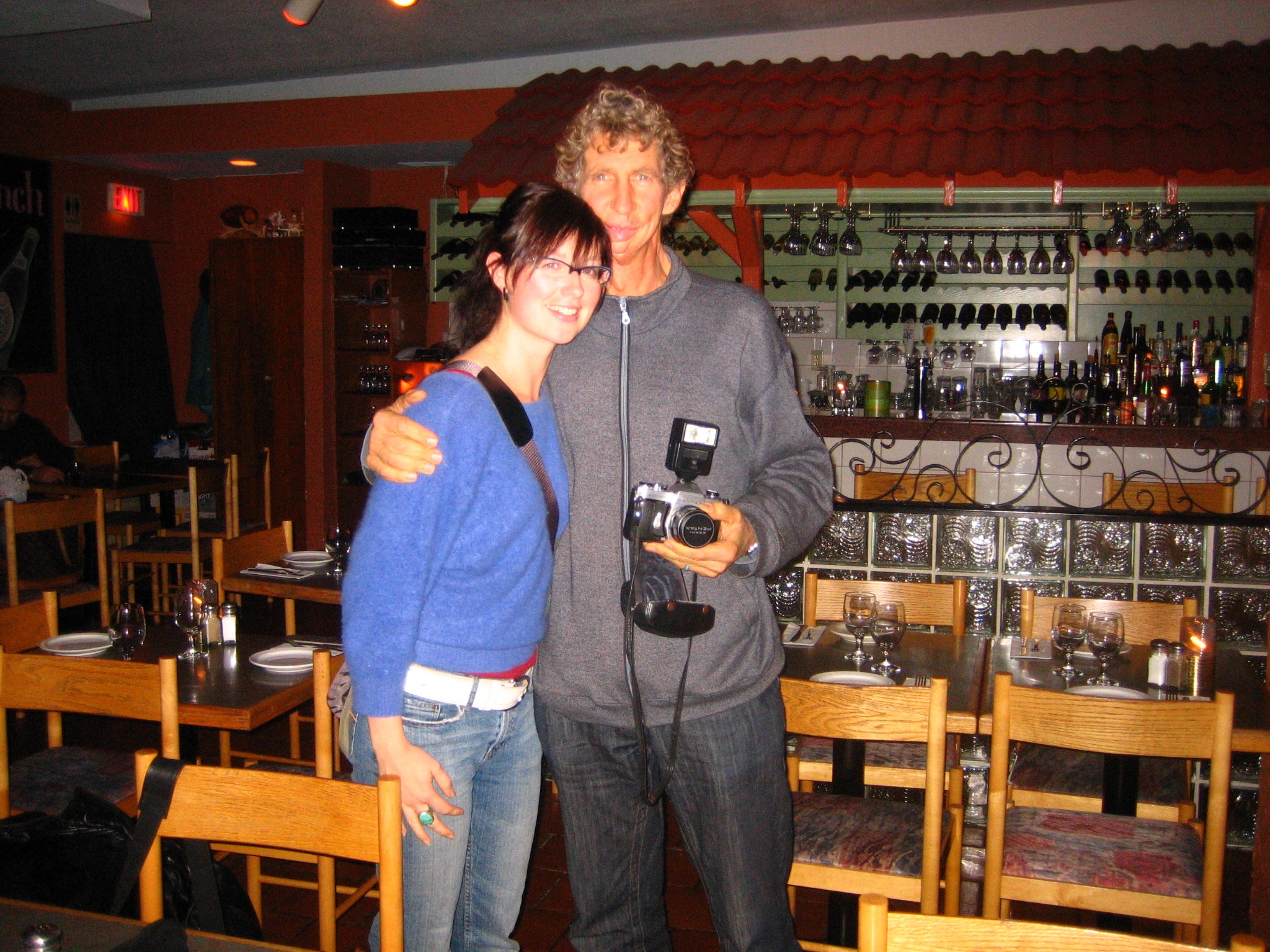 woman in blue sweater and jeans standing next to her father with his arm around her, he's holding a camera with a flash installed on the top in a restaurant
