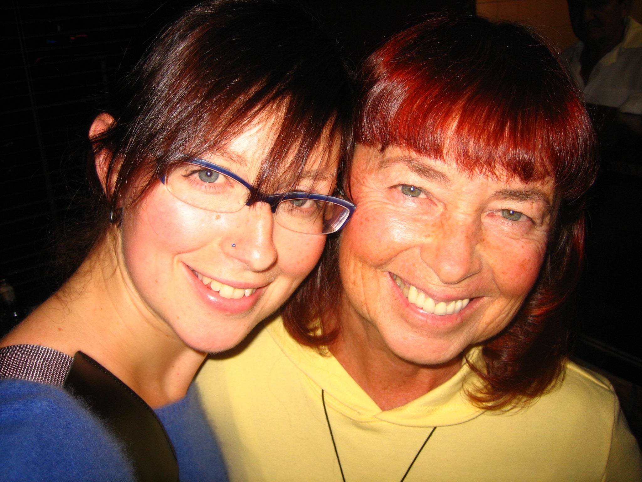 close up photo of woman wearing glasses in blue fuzzy sweater next to her mother with red hair and a yellow top, both are smiling broadly and beautifully