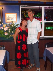 sandi wearing black and red print dress and bill in sandals, blue pants and mostly white shirt standing in a colourful restaurant