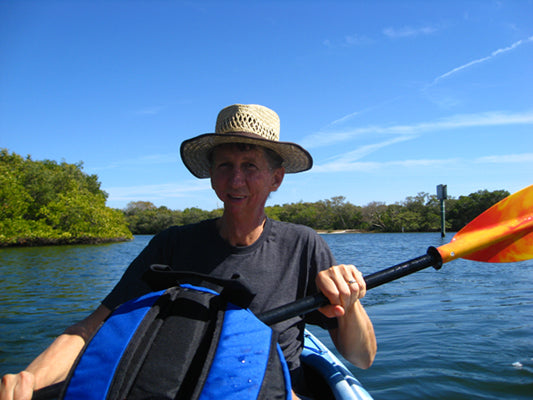 smiling man in a black t shirt and wearing straw hat and holding a paddle sitting in a kayak in the water on a sunny summer day