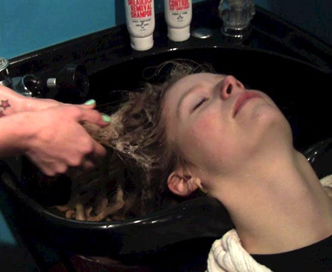 woman laying back in salon sink with her eyes closed as a stylist's hands are seen shampooing her dreadlocks