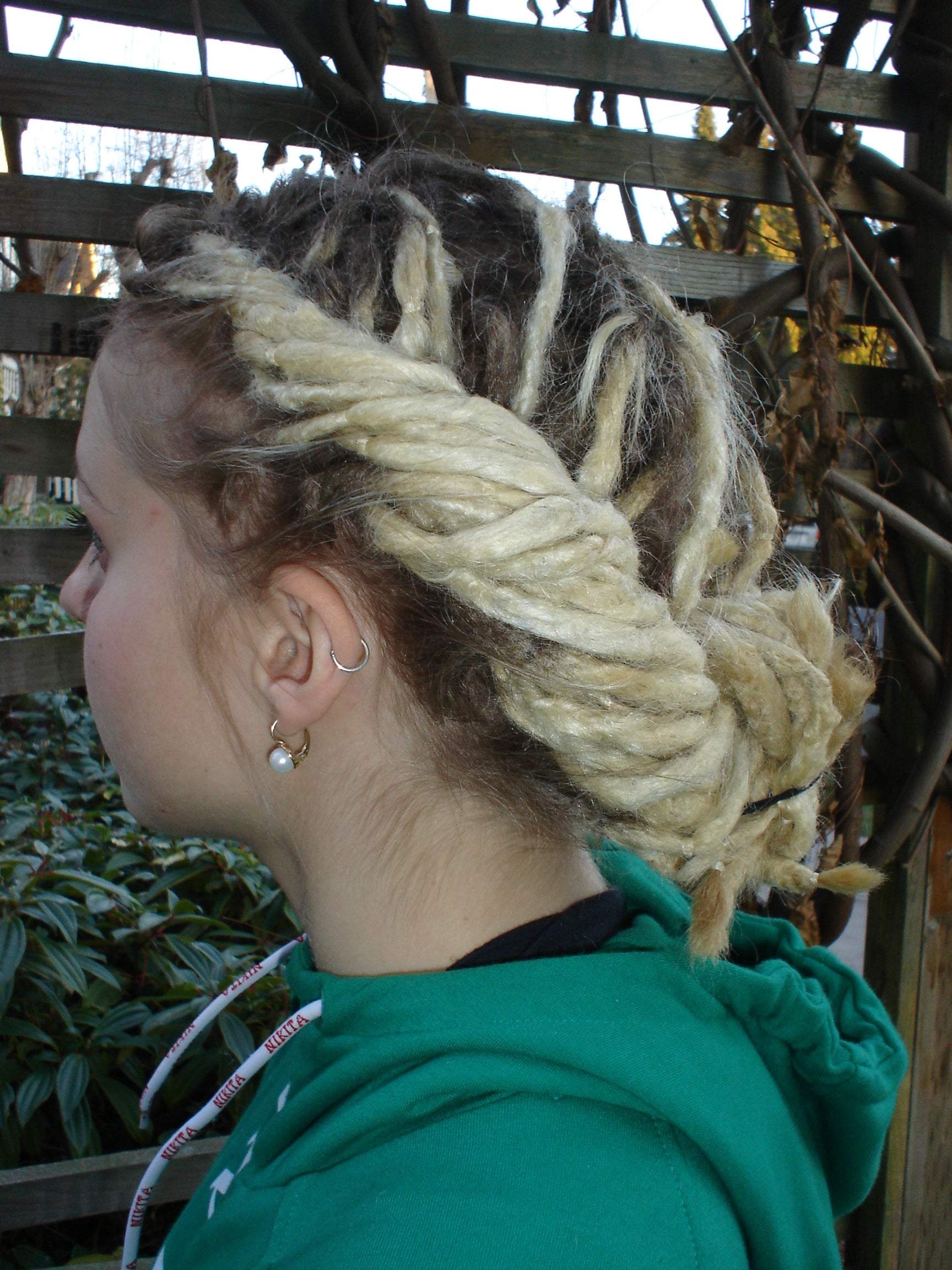 woman with light blonde coloured dreadlocks standing in front of a trellis showing left hand rear view of french draids hairstyle