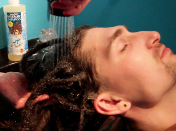man leaning back in salon sink with his eyes closed against a blue background having his dreadlocks rinsed