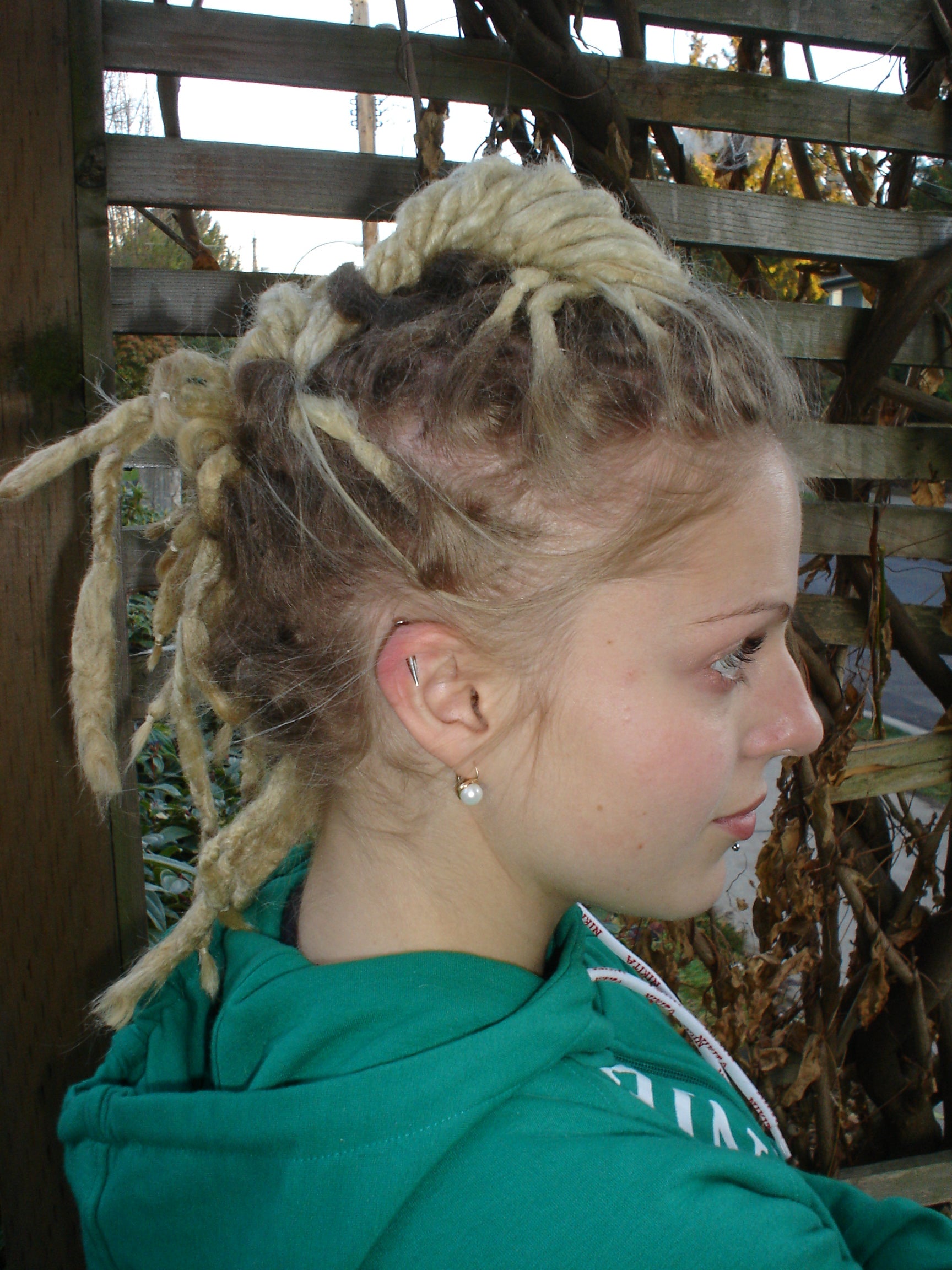 side view of dreadlock mohawk hairstyle modelled by young woman with dyed blonde dreadlocks wearing green shirt in front of arbor