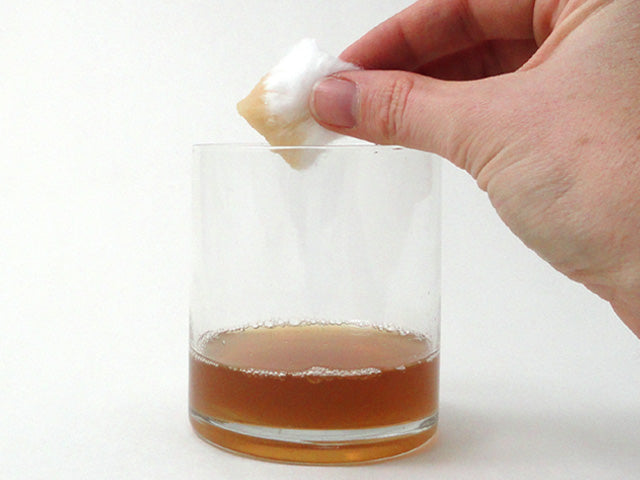 hand holding a cotton ball above a clear glass of dark amber rosemary infusion