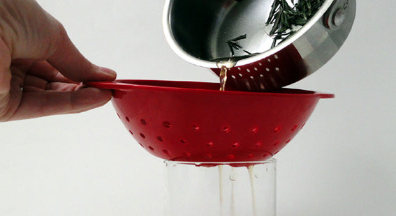 hand holding edge of red plastic strainer while infusion from a small pot of boiled rosemary is strained through it