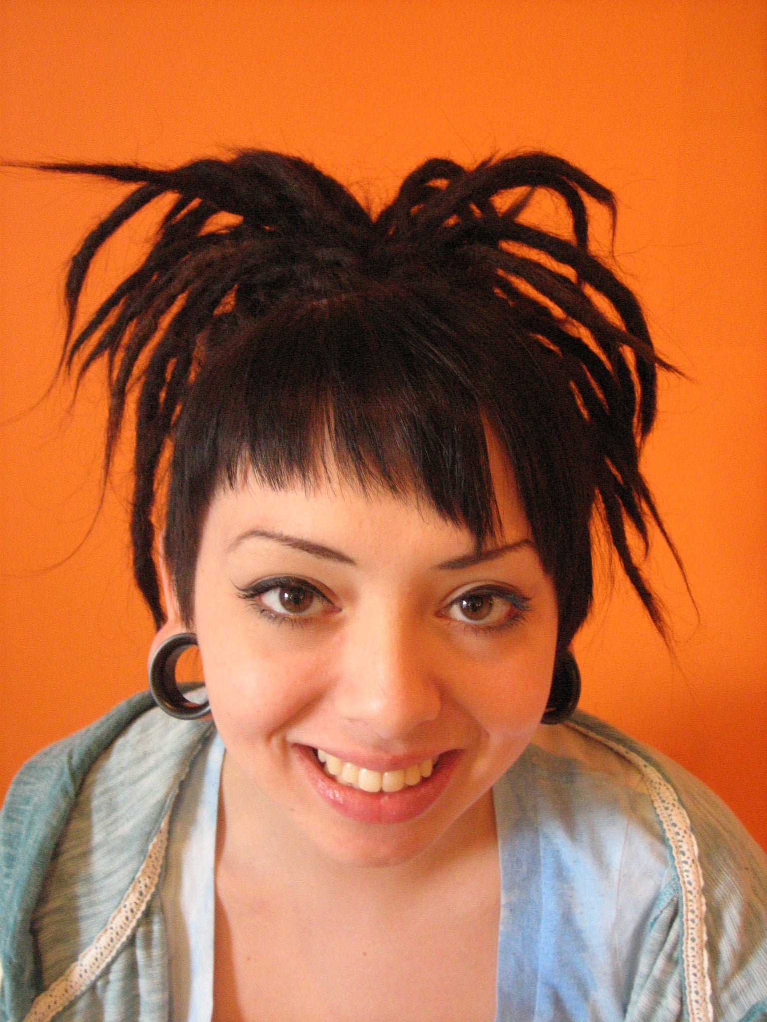 smiling woman with large earring flesh tunnels and dark brown dreadlocks against an orange background with bangs and knotted dreadlock do