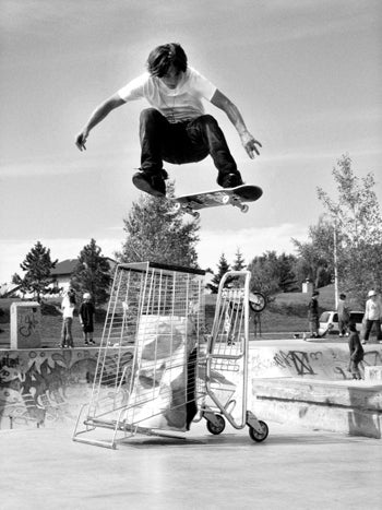 black and white photo of a young man performing quite impressive skateboard jump over an upturned shopping cart