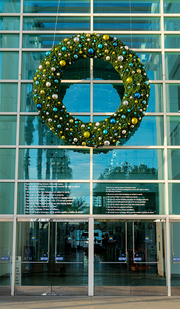 A giant artificial Christmas wreath decorated with light green, turquoise, blue, and silver ornaments hangs on a giant entryway made of windows.