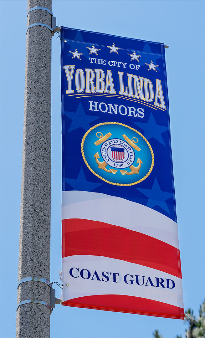 Close-up of a vertical patriotic banner on a lamp post in Yorba Linda, featuring the United States Coast Guard emblem. The banner is blue with white stars at the top and red and white stripes at the bottom, reading &quot;The City of Yorba Linda Honors Coast Guard.&quot;