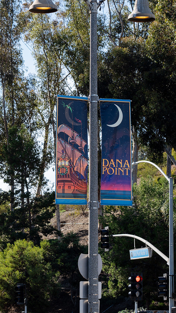 Two banners hanging from a streetlamp during the day. The banners feature an illustrated ship under a crescent moon with the text &quot;Dana Point.&quot; The background includes trees and street signs.