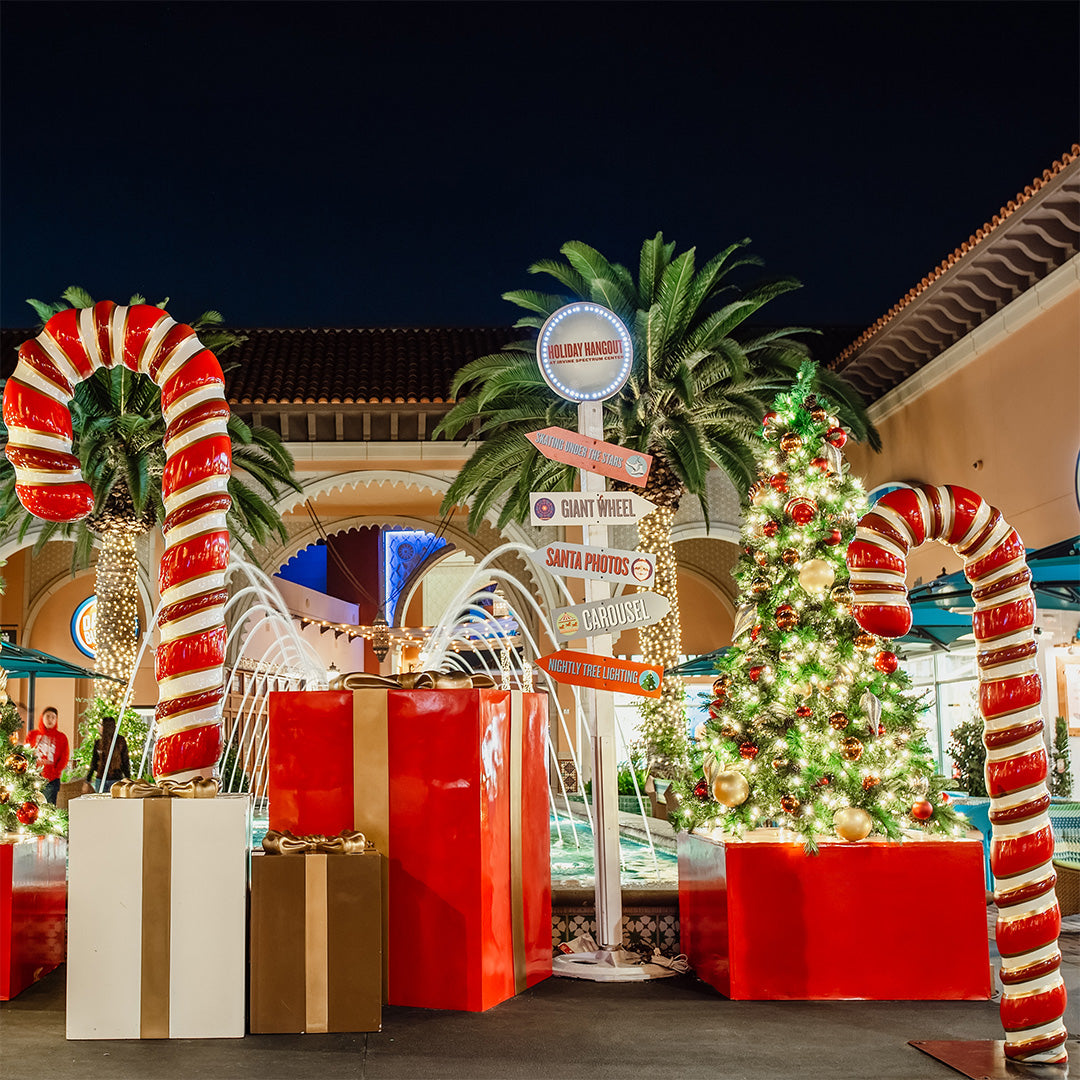 Giant fiberglass props including candy canes and gift boxes are staged alongside a decorated Christmas tree and navigational signage, indicating the direction of various holiday photo ops.