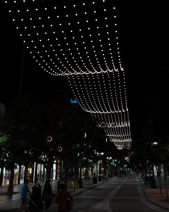 A curtain of warm white globe light bulbs is draped over an outdoor shopping center walkway.