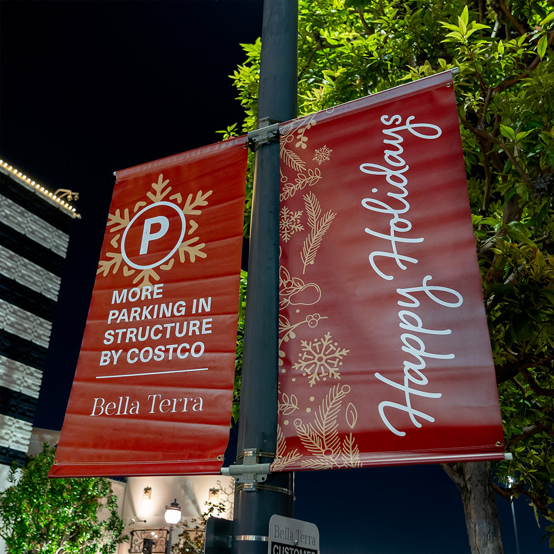 Two red holiday banners displayed on a street lamp at night. One banner reads "More Parking in Structure by Costco" with a large "P" symbol, and the other says "Happy Holidays" with festive designs. The banners are set against a backdrop of a tree and buildings decorated with lights.