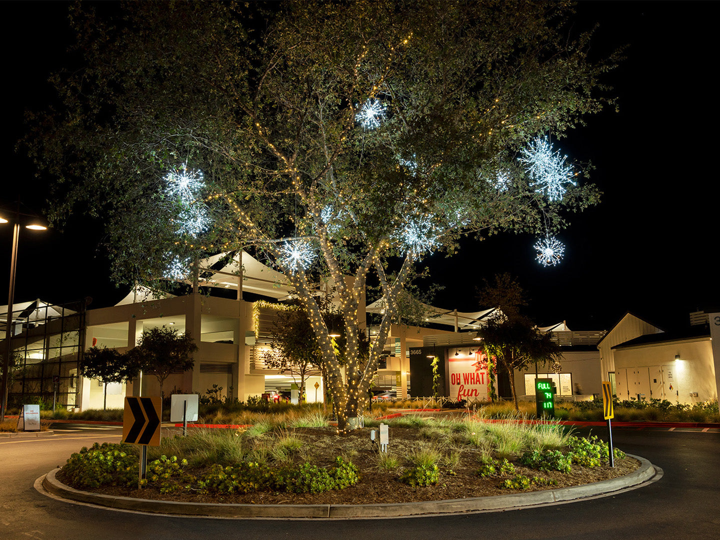 A tree in the middle of a shopping center driveway is decorated with warm white mini lights and cool white starburst decorations.