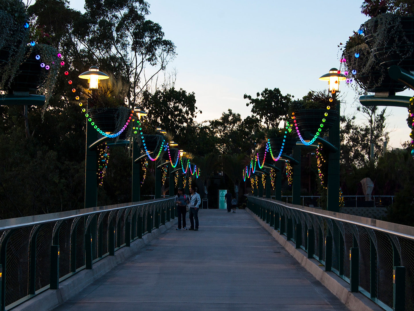 A bridge is decorated with one strand of rainbow lights draped on both sides of the walkway.
