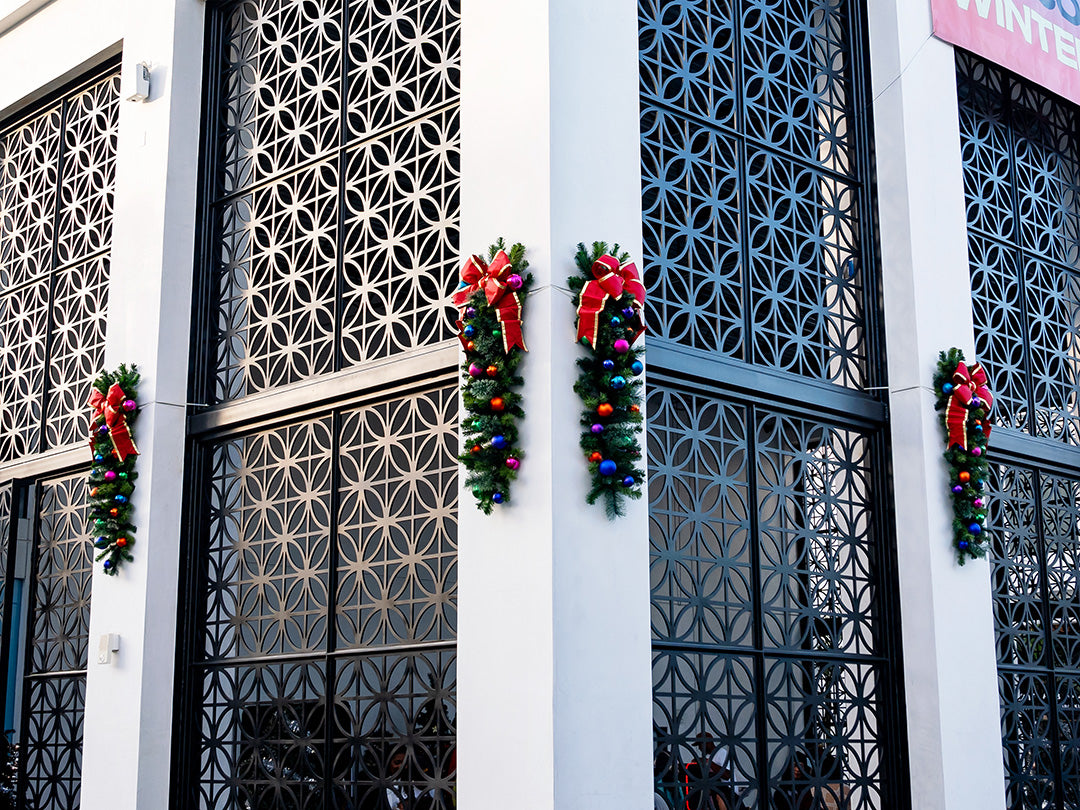A close-up of a Christmas garland with red and silver ornaments and a red bow, attached to the exterior wall of a building.