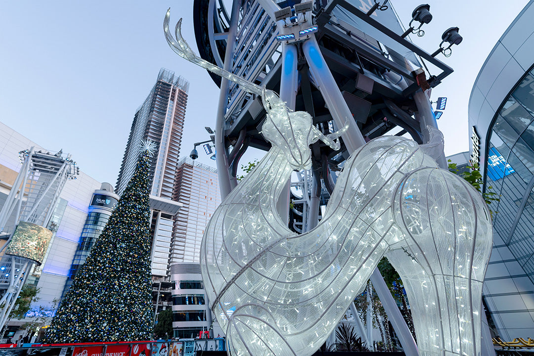 A large, illuminated deer statue graces a city square, with a brightly decorated Christmas tree behind it.