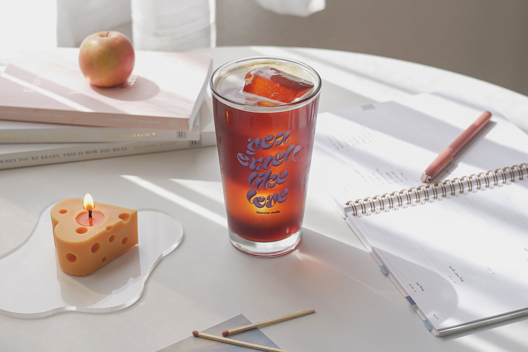 Vaso de café helado, libros, vela de queso en forma de corazón sobre un posavasos acrílico y un diario sobre la mesa