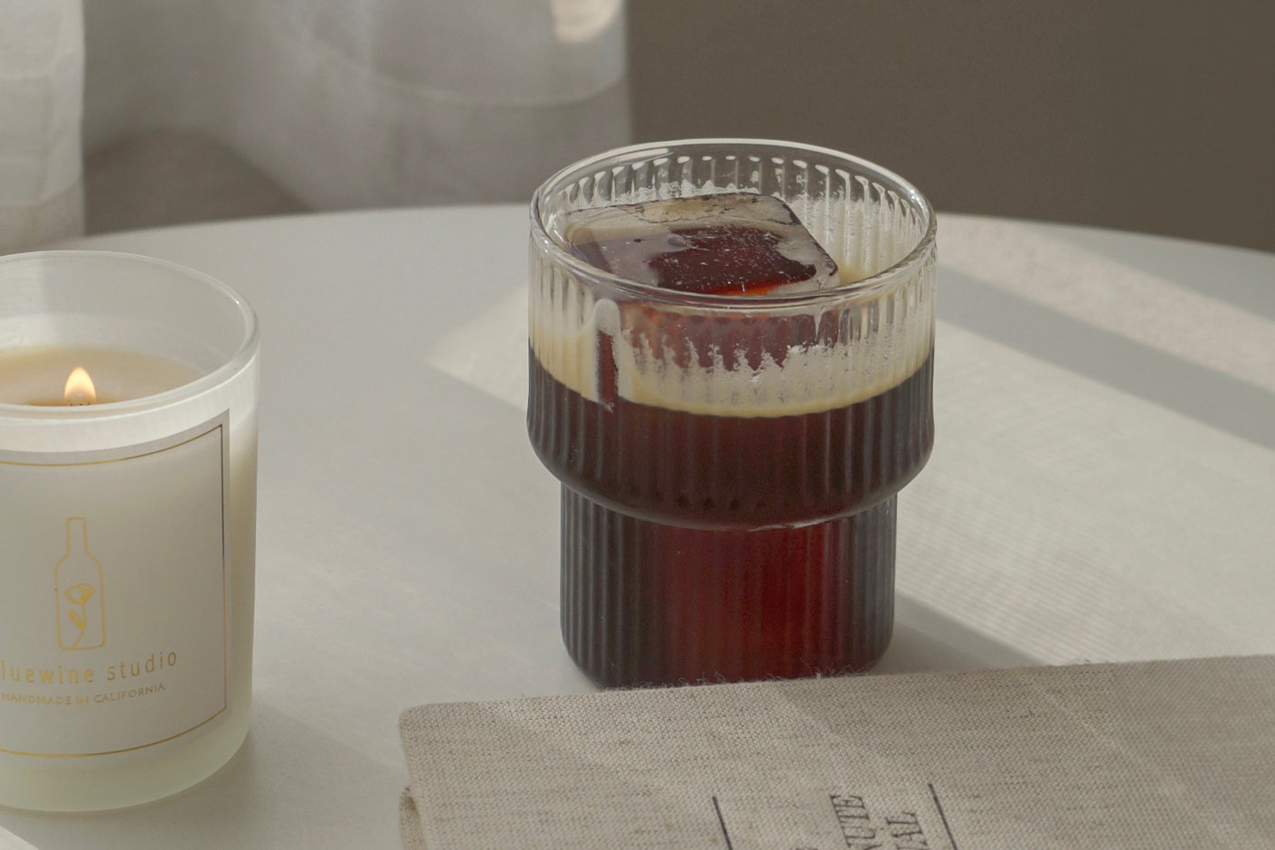 불이 붙은 5온스 간장 양초, 골이 있는 유리에 담긴 아이스 커피, 흰색 원형 테이블 위에 놓인 5분 일기
