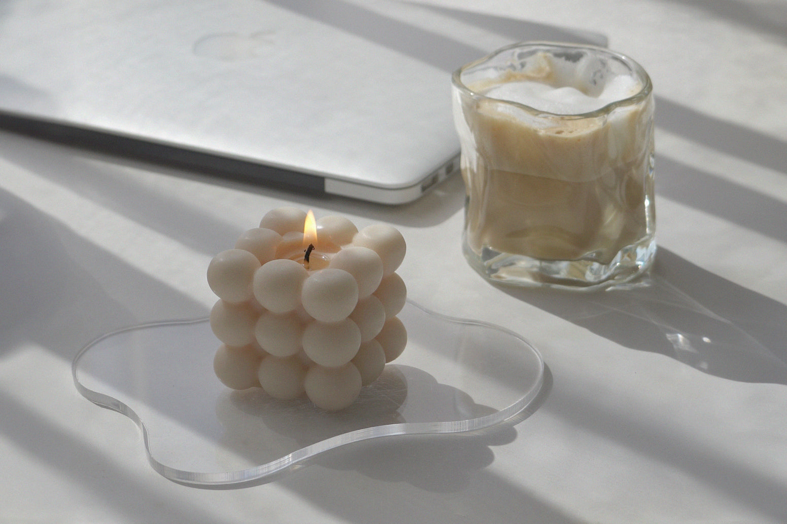 una vela cúbica de color beige encendida sobre un posavasos acrílico transparente, irregular y ondulado y un café con leche helado con crema encima en un vaso irregular de 6 oz