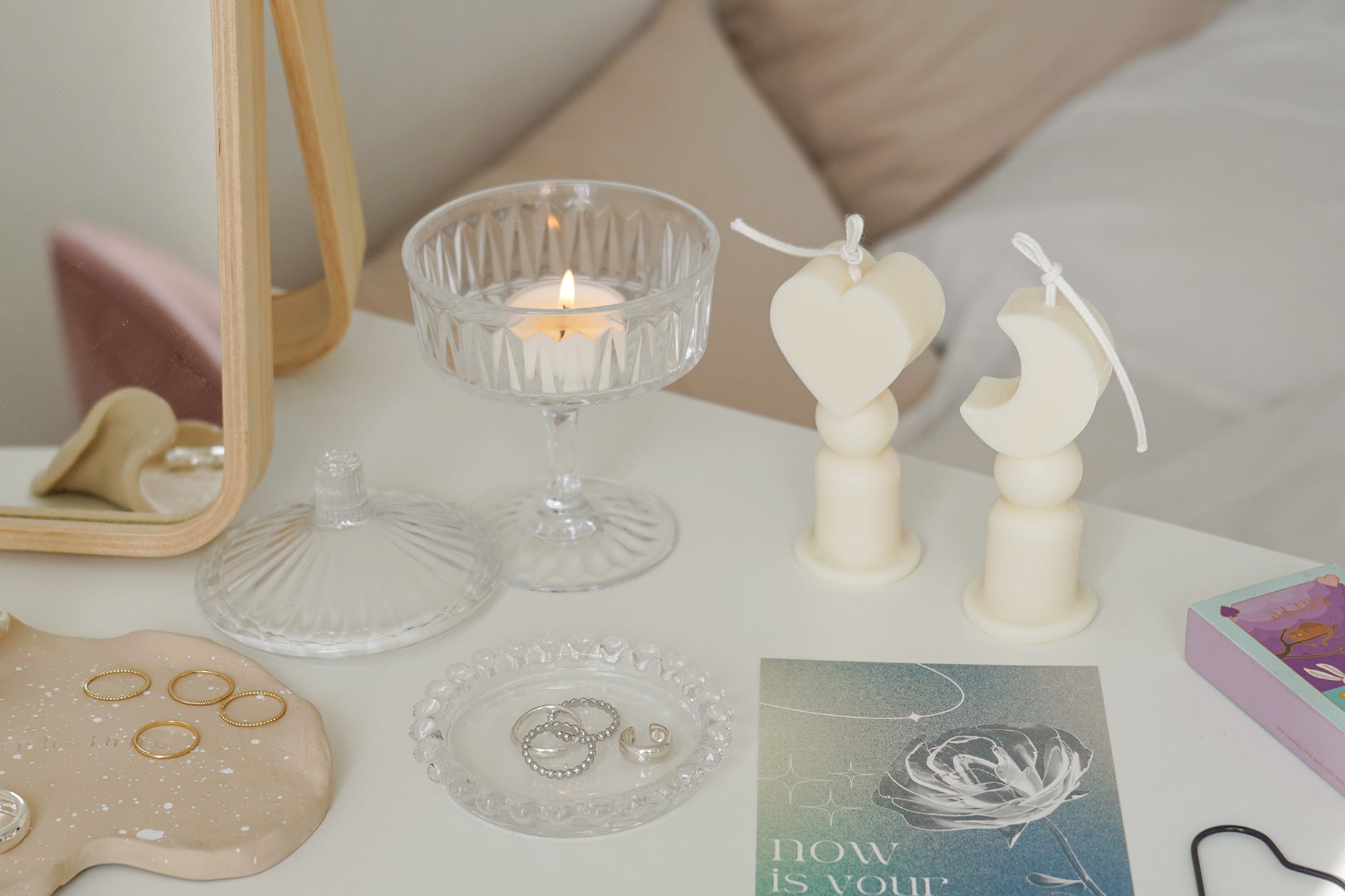 Heart shape candle, crescent moon shape candles, a lit tealight candle in a glass coupe, blue postcard, match box, black heart shape wick dipper, and silver rings on clear beaded tray are place on white drawer. The arrangement exudes minimal aesthetic dreamy vibes