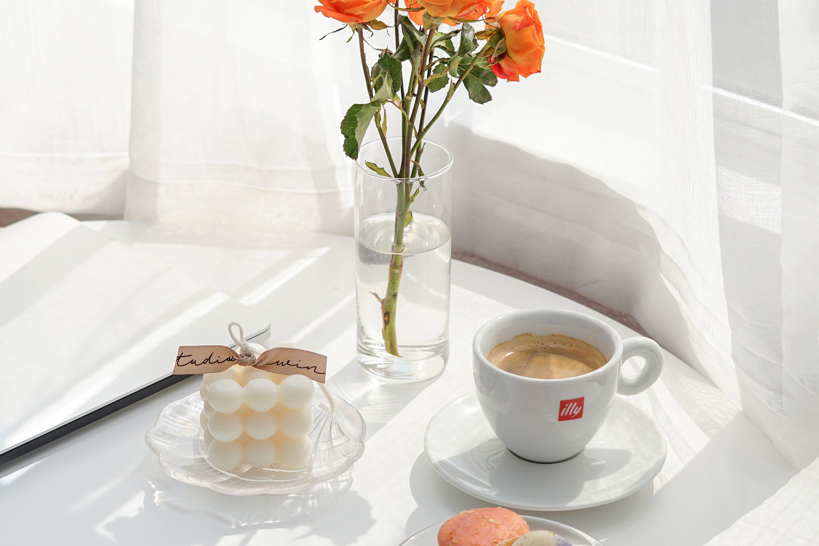 투명한 꽃병에 있는 오렌지색 장미, 홀로그램 쉘 트레이에 있는 큐브 캔들, 커피 한 잔, 흰색 원형 테이블에 있는 노트북