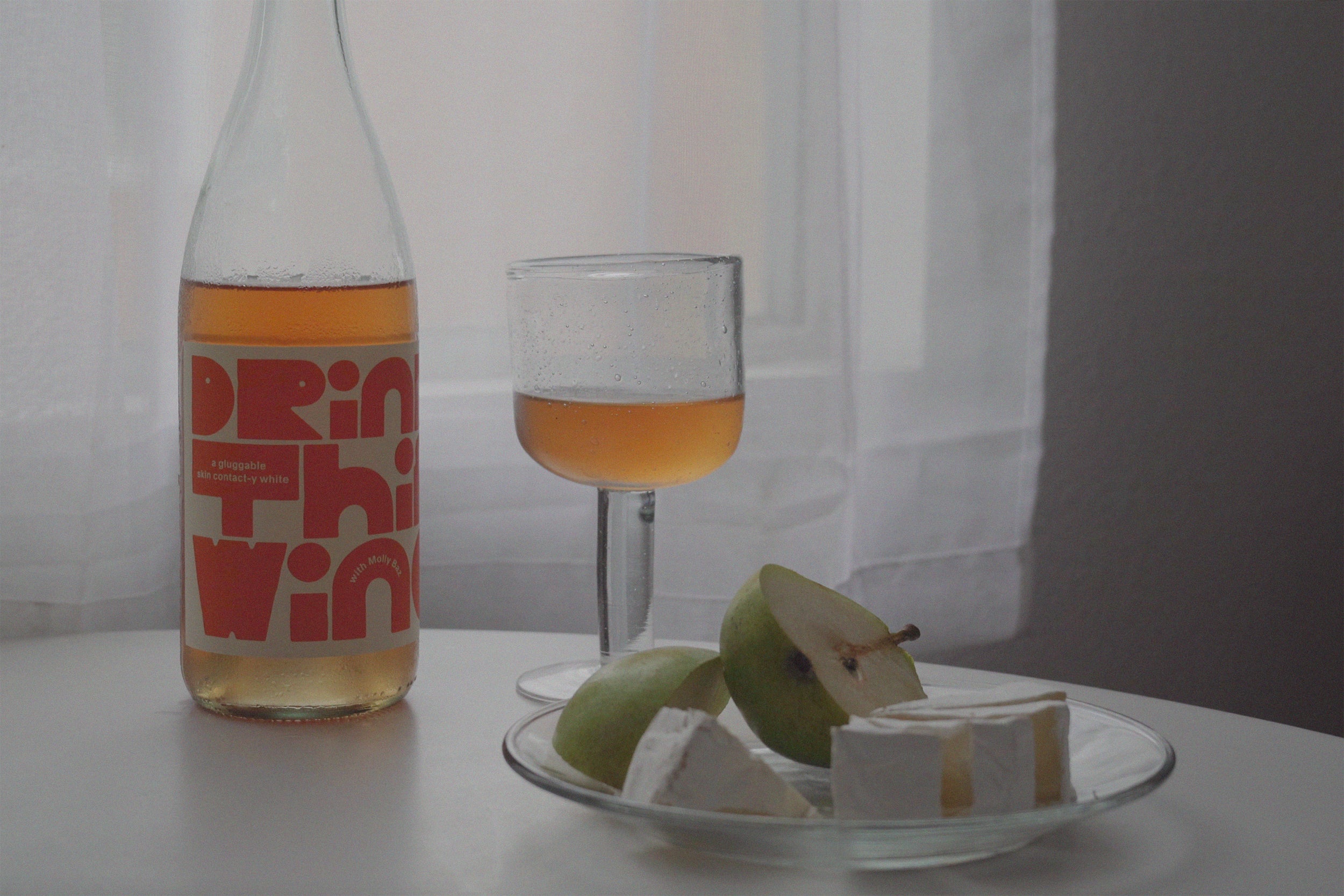 una botella de vino de naranja, una copa de vino de naranja y rodajas de queso brie y rodajas de pera en un plato transparente colocado sobre una mesa redonda blanca